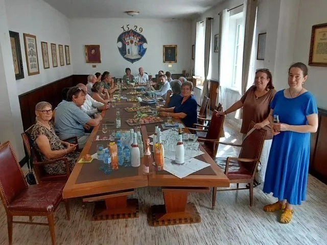 Gäste sitzen an einem festlich gedeckten Tisch in einem Speisesaal, zwei Frauen stehen daneben.
