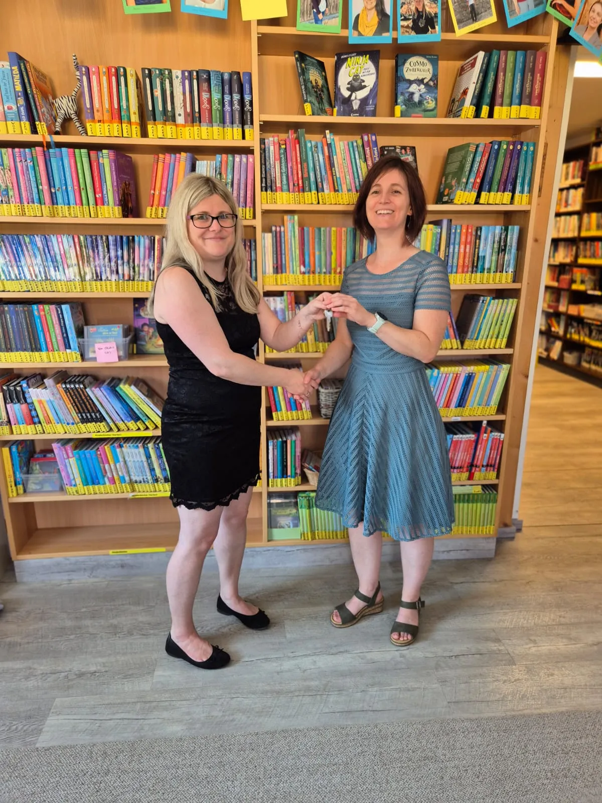 Zwei Frauen übergeben sich einen Schlüssel vor einem Bücherregal in einer Bibliothek