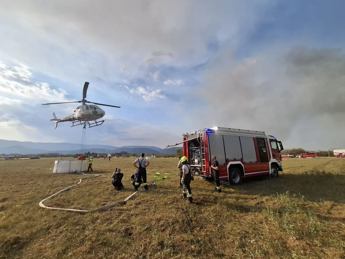 Hubschrauber im Einsatz über einem Feld mit Feuerwehr und Löschwasserbehälter