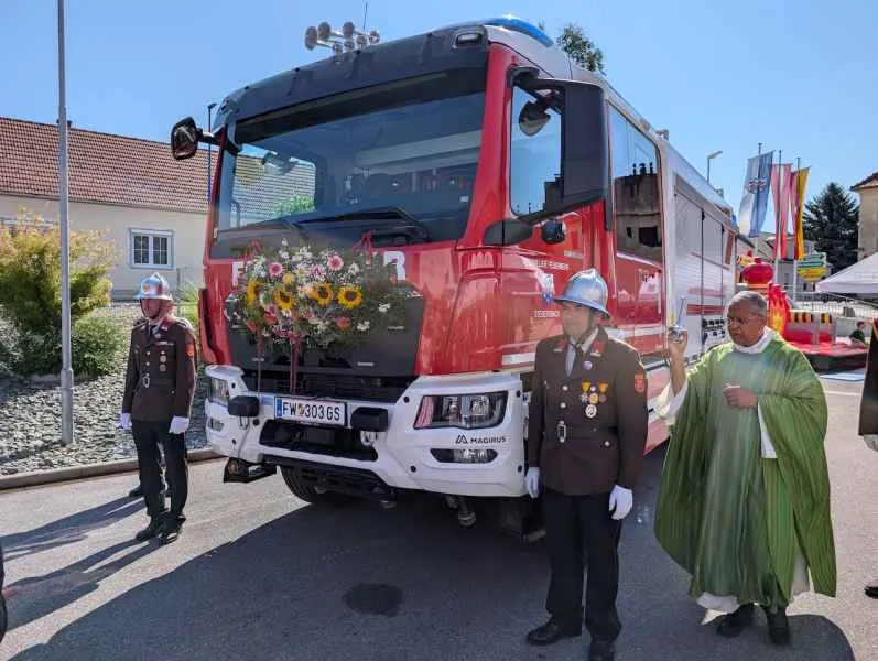 Feuerwehrfest in Niederösterreich: Zwei Feuerwehrmänner und ein Geistlicher stehen vor einem geschmückten Feuerwehrauto.