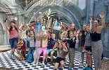 Kindergruppe winkt fröhlich vor einer bunten Kulisse mit Schachbrettboden.