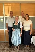 Vier Personen in Tracht und Anzug stehen in einem Raum mit gemustertem Boden und Glaswänden.