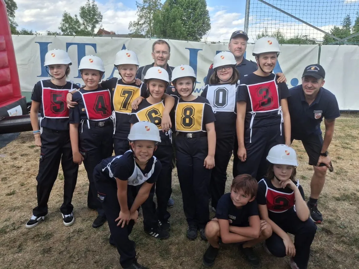 Jugendfeuerwehr-Wettkampf mit Kindern und Betreuern in Uniformen und Helmen vor einer Fahne