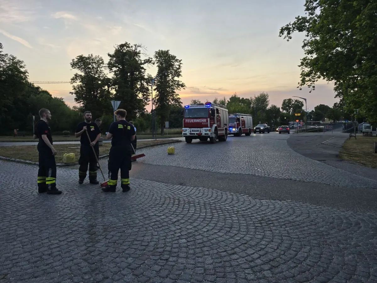 Feuerwehrleute mit Besen stehen vor Feuerwehrfahrzeugen auf einem Kopfsteinpflasterplatz.