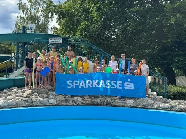 Kinder und Erwachsene mit Schwimmhilfen vor Sparkassen-Banner im Freibad