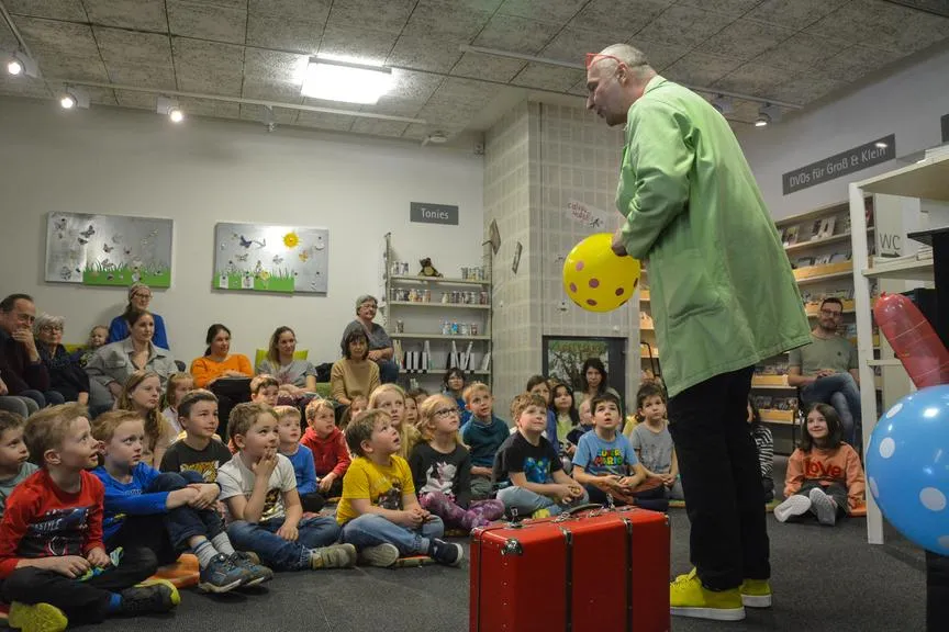 Mitmacher-Zauberer mit gelbem Ballon und rotem Koffer vor Kindern im Gemeindesaal