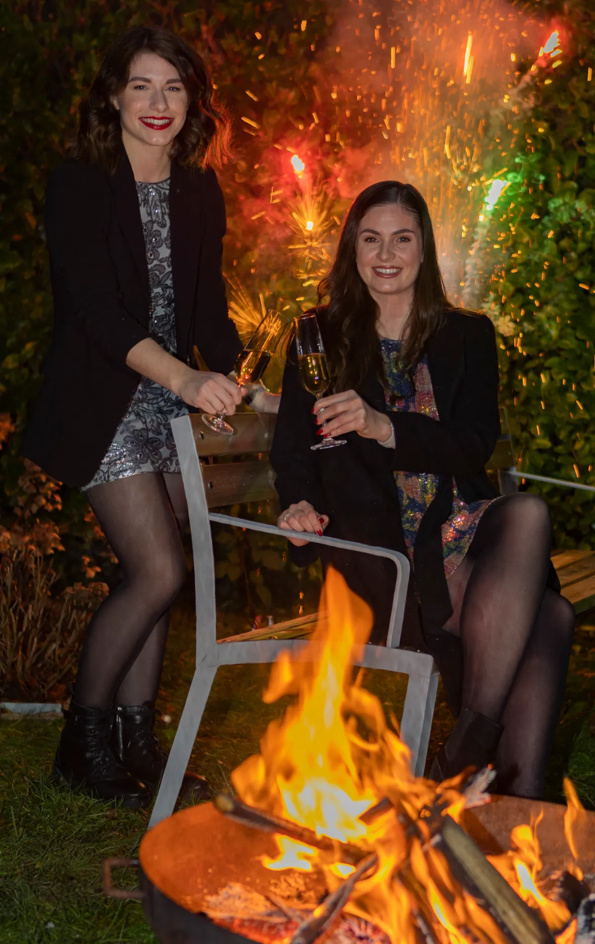 Zwei Frauen stoßen mit Sektgläsern an, Feuerwerk und Lagerfeuer im Garten
