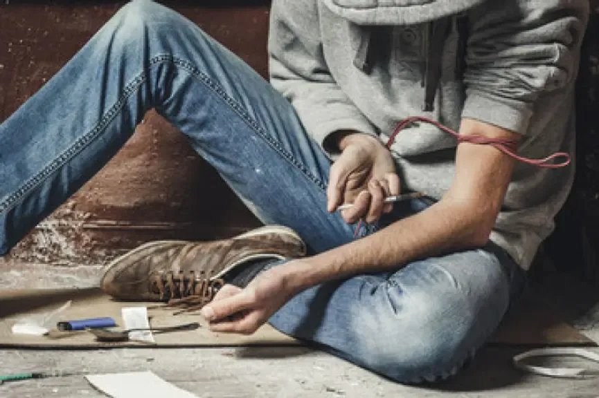Junge Person mit Spritze und Tourniquet im Arm, auf dem Boden sitzend.