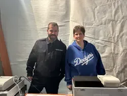 Mann und Frau stehen hinter einem Tisch mit Registrierkassen in einem provisorisch eingerichteten Raum.