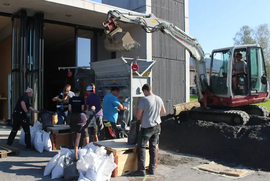 Bagger leert Schotter in eine Mischmaschine, während Männer in Österreich Säcke füllen.