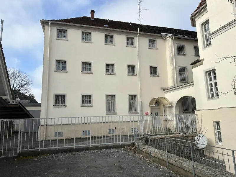 Gitter vor Fenstern an einem hellen Gebäude mit Ziegeldach, im Hintergrund ein Baum