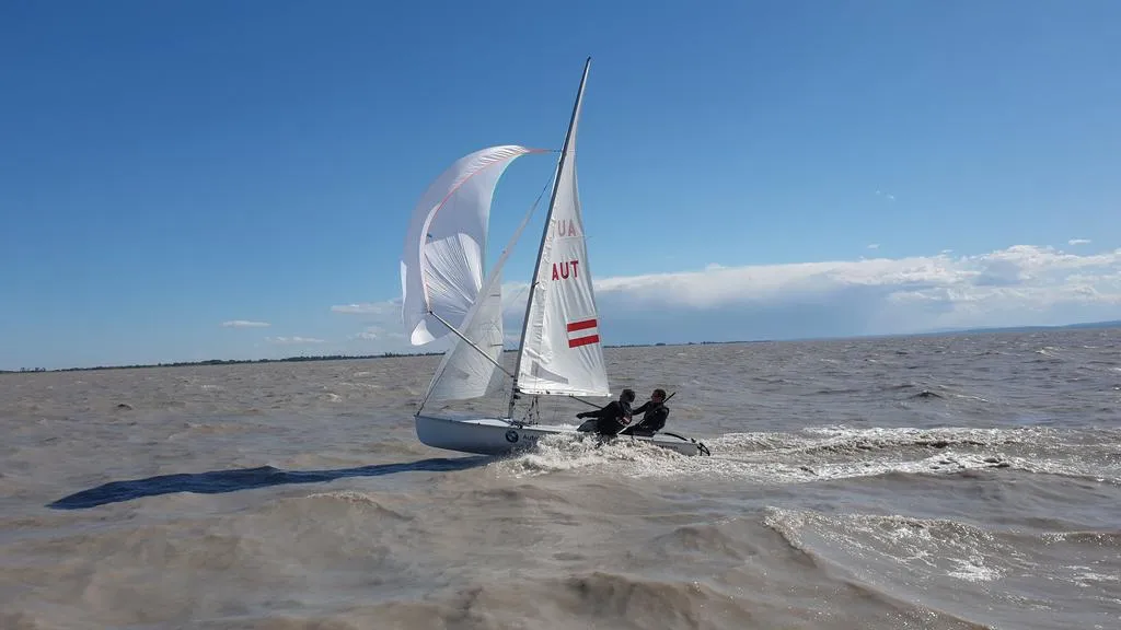 Segelboot mit zwei Personen auf stürmischer See vor flacher Küstenlinie