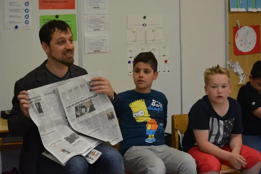Lehrer hält Zeitung, zeigt Kindern in einem Klassenzimmer in Österreich Artikel.