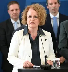 Beate Meinl-Reisinger vor Mikrofon in Wien, im Hintergrund Männer in Anzügen.