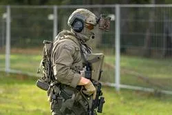 Soldat mit Helm und Ausrüstung im Freien, mit Zaun im Hintergrund.