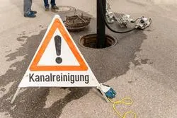 Kanalreinigung: Warnschild und offener Schacht mit Ausrüstung auf einem nassen Asphalt.