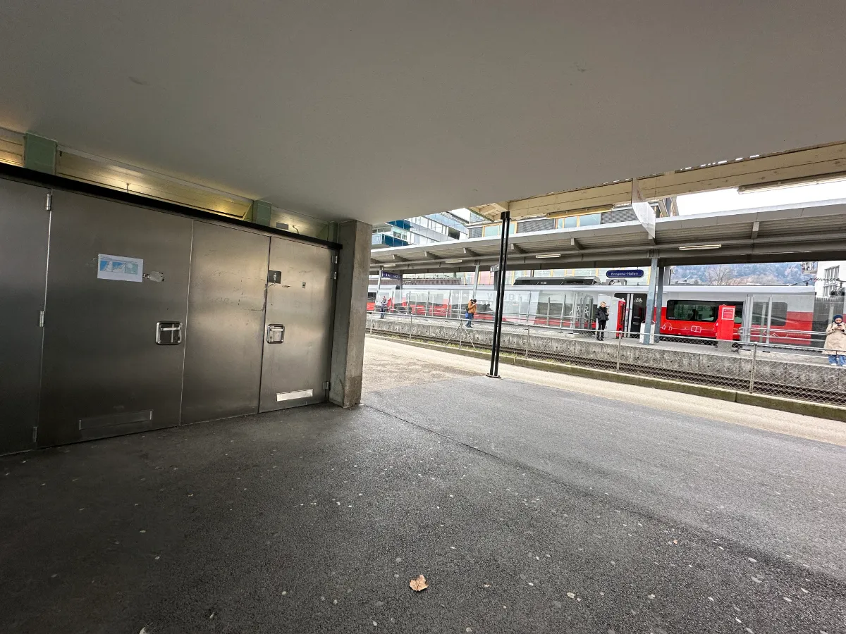 Bahnhof Bregenz-Hafen mit roten Zügen und wartenden Personen unter einem Vordach