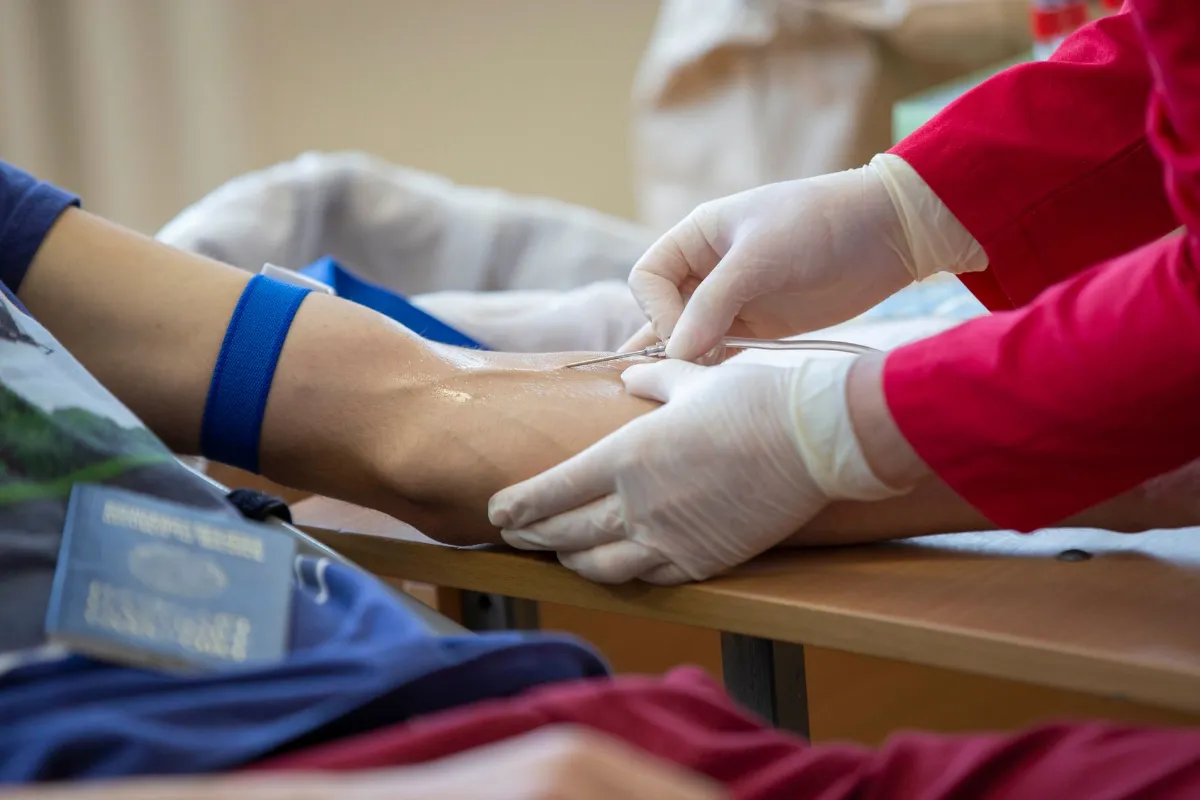 Blutspende: Sanitäterin mit Handschuhen legt Nadel am Arm eines Spenders an