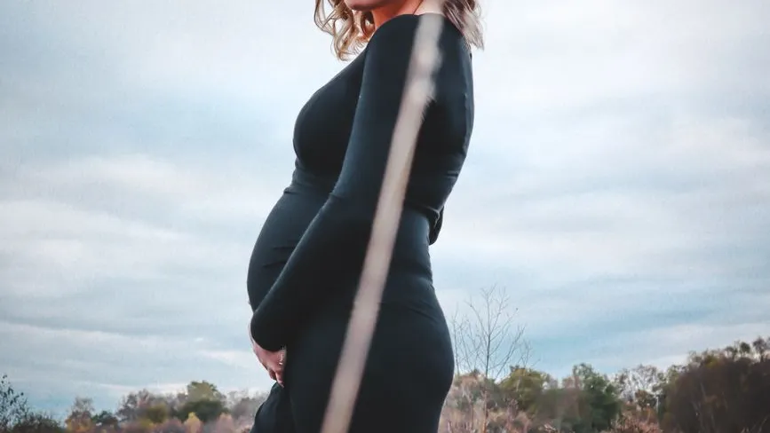 Schwangerschaft im Freien, Frau in schwarzem Kleid mit sichtbarem Bauch vor bewölktem Himmel