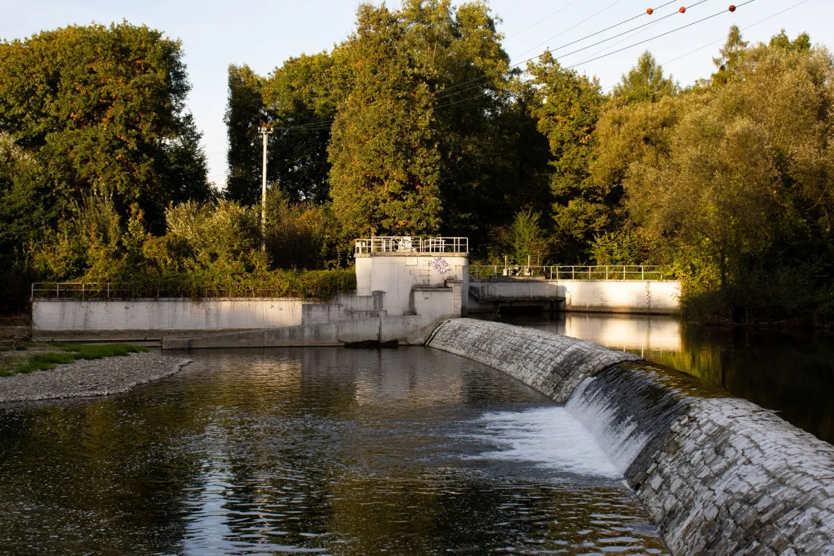 Staustufe mit Steinböschung und Betonbauwerk im bewaldeten Uferbereich eines Flusses