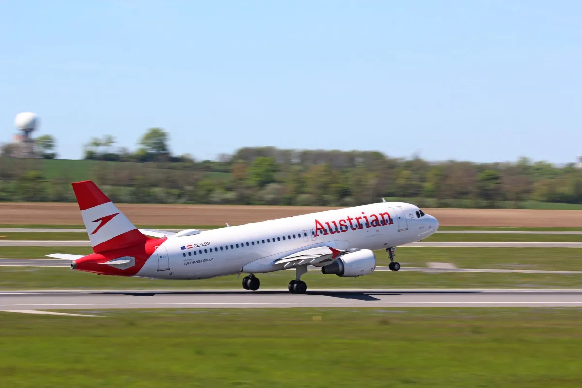 Austrian Airlines Flugzeug hebt auf Rollfeld ab, mit Bäumen und Himmel im Hintergrund.