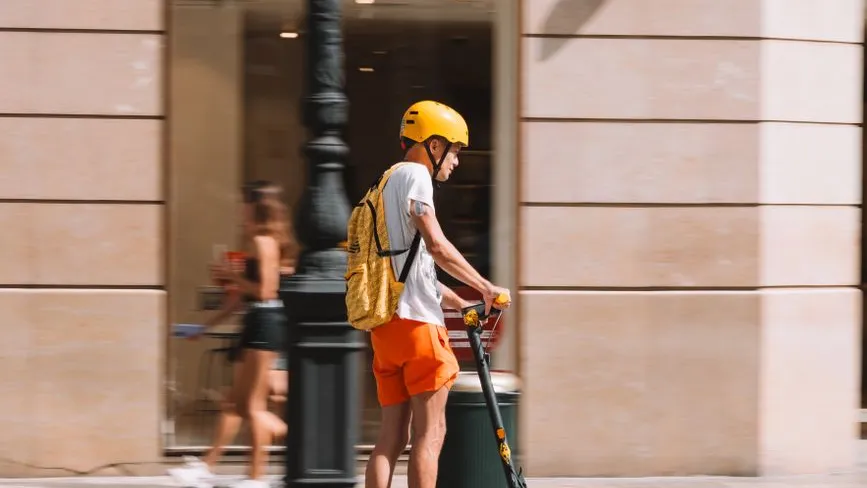 Elektrorollerfahrer mit gelbem Helm und Rucksack fährt in Wien vor einem Geschäft.