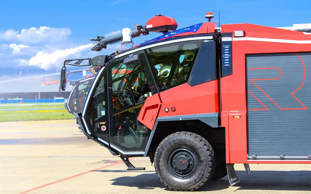Löschfahrzeug mit ausgefahrenem Wasserwerfer auf einem Flugfeld, besetzt mit Feuerwehrmännern.