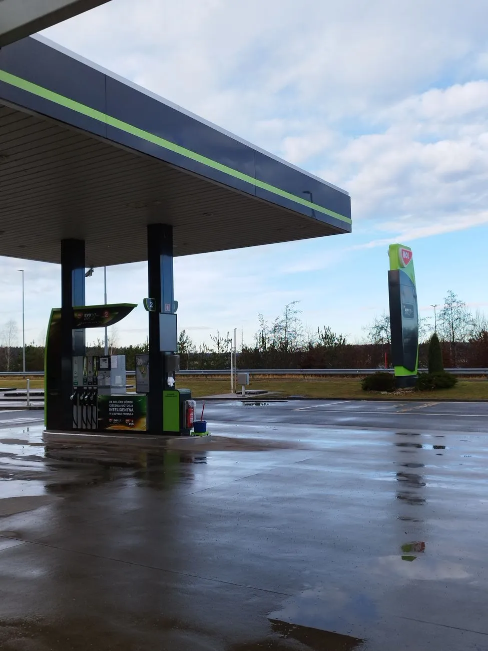Tankstelle mit Zapfsäulen und Werbesäule bei bewölktem Himmel, nasser Boden