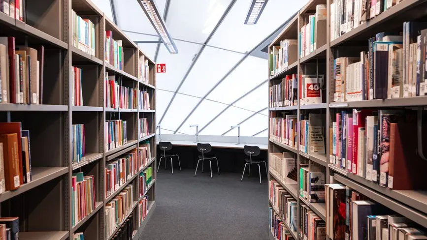 Bücherregale in einer Bibliothek mit Arbeitsplätzen und Stühlen im Hintergrund