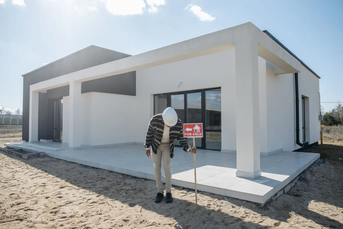 Mann mit Bauhelm und "For Sale"-Schild vor neuem Einfamilienhaus