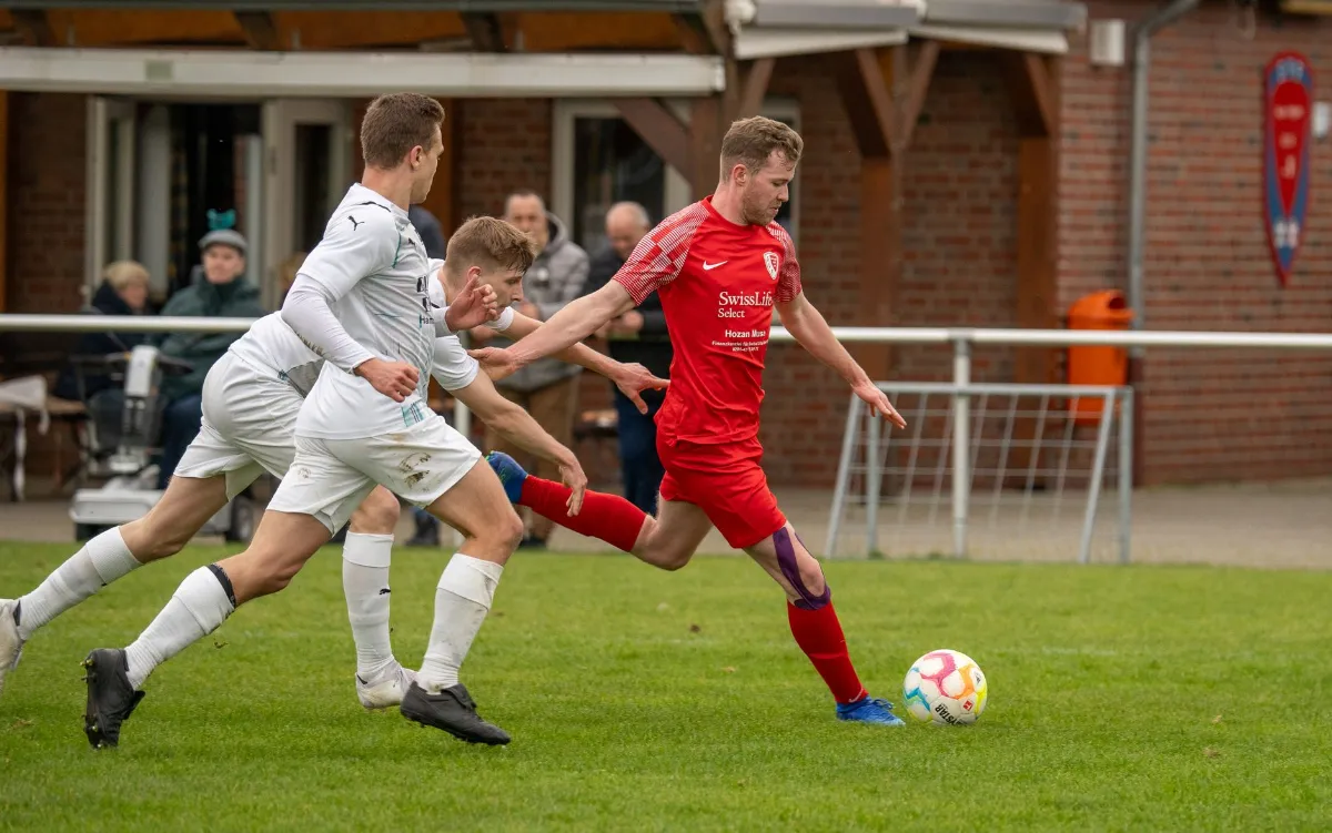 Fußballspieler in Rot schießt auf Ball, zwei Spieler in Weiß verteidigen