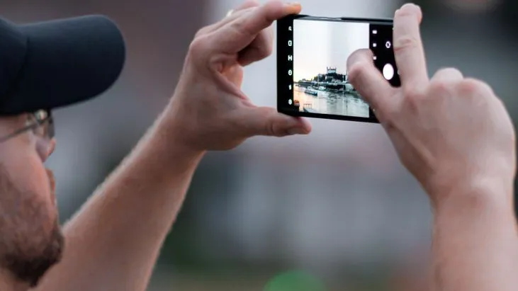 Mann mit Kappe fotografiert mit Smartphone eine Stadtansicht mit Fluss und Booten.