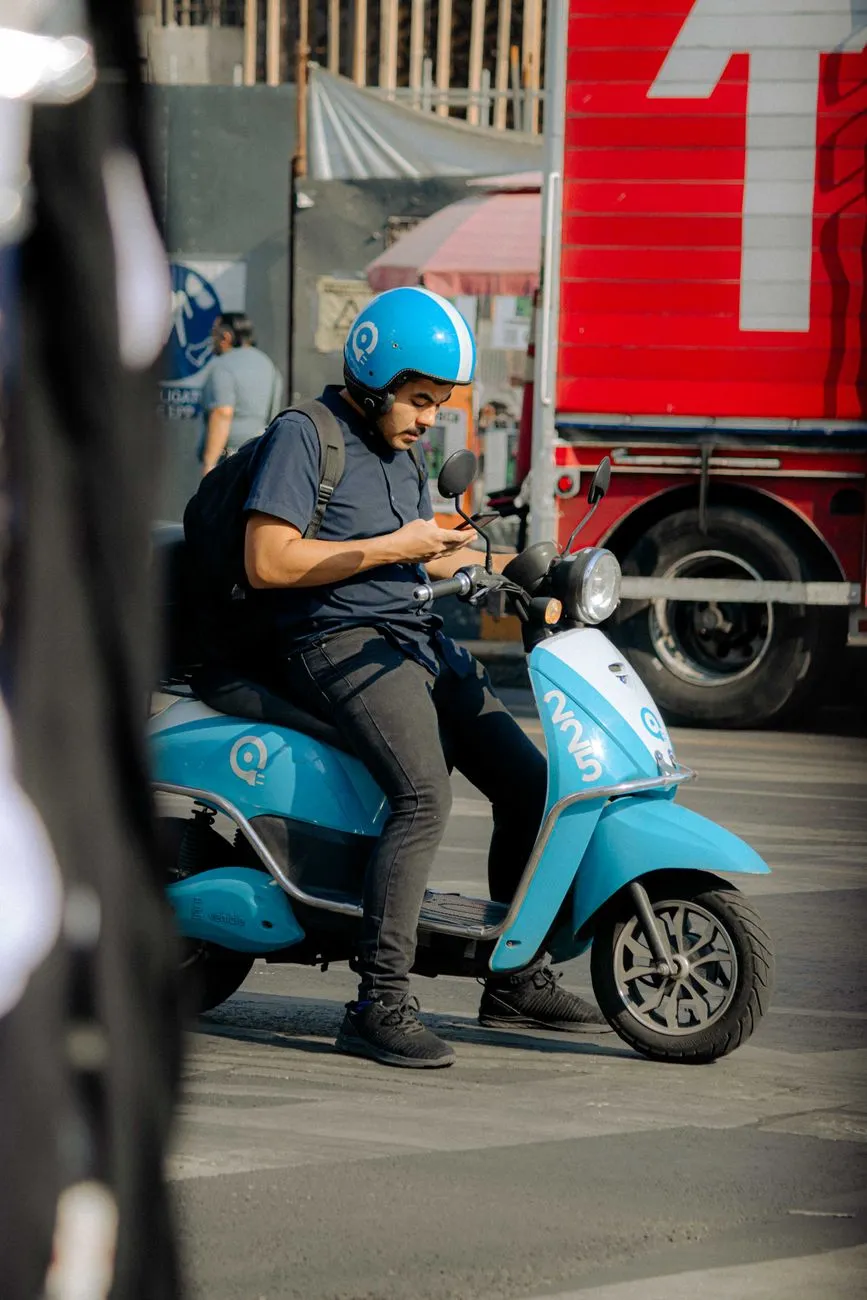 Rollerfahrer mit blauem Helm und Rucksack schaut aufs Handy, davor rote LKW-Plane