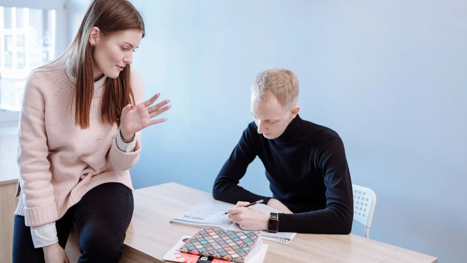 Junge Frau mit hellrosa Pullover gestikuliert, Mann mit schwarzem Rollkragenpullover schreibt am Tisch