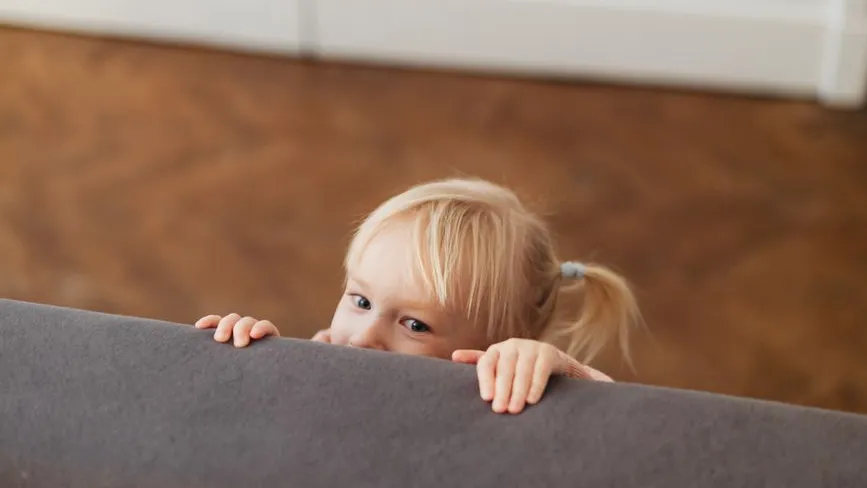 Kleines Mädchen mit blonden Zöpfen lugt hinter einer grauen Couch hervor.