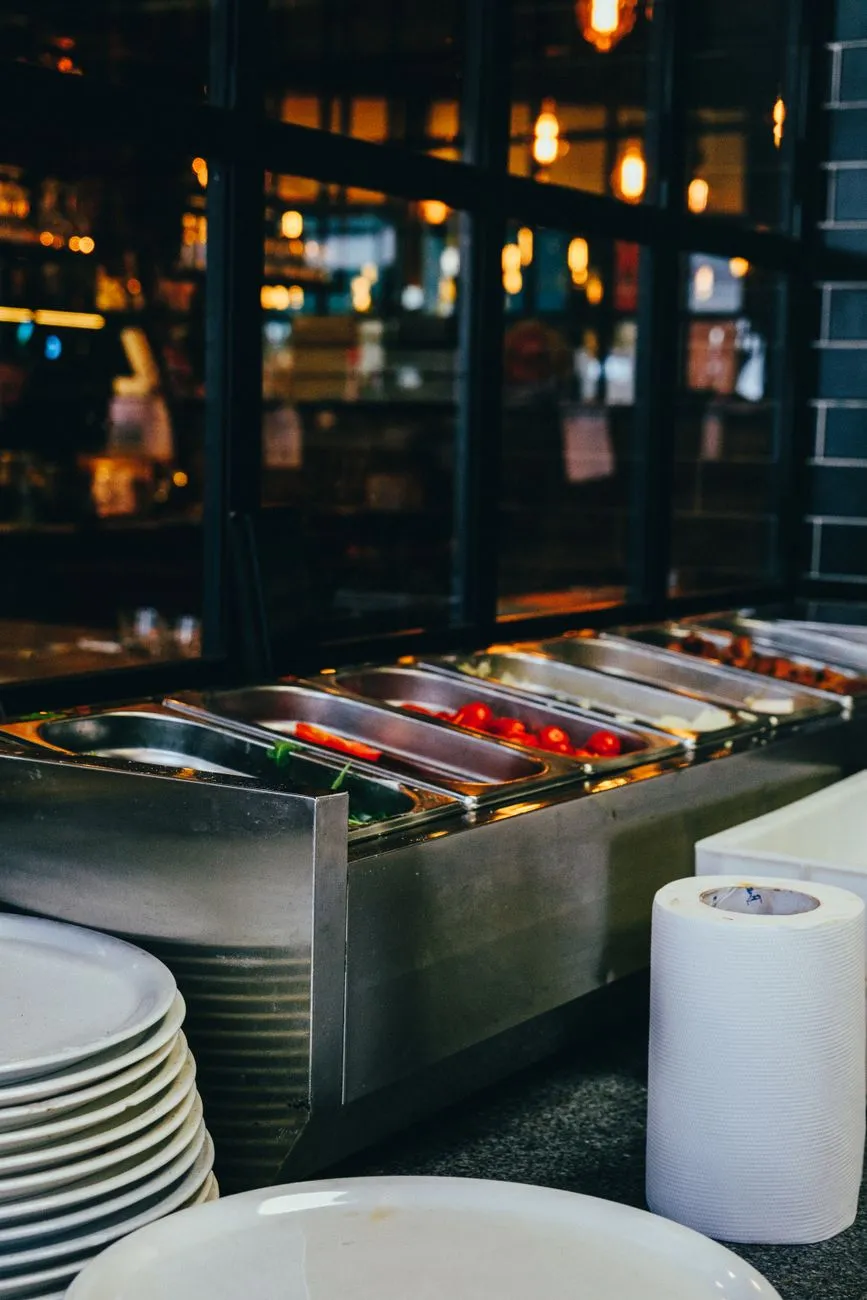 Buffet mit verschiedenen Speisen in Edelstahlschalen, Tellerstapel und Küchenrolle