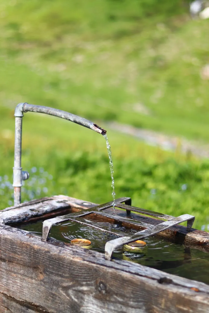 Trinkbrunnen aus Holz mit Metallrohr, aus dem Wasser in einen Trog mit Münzen fließt.
