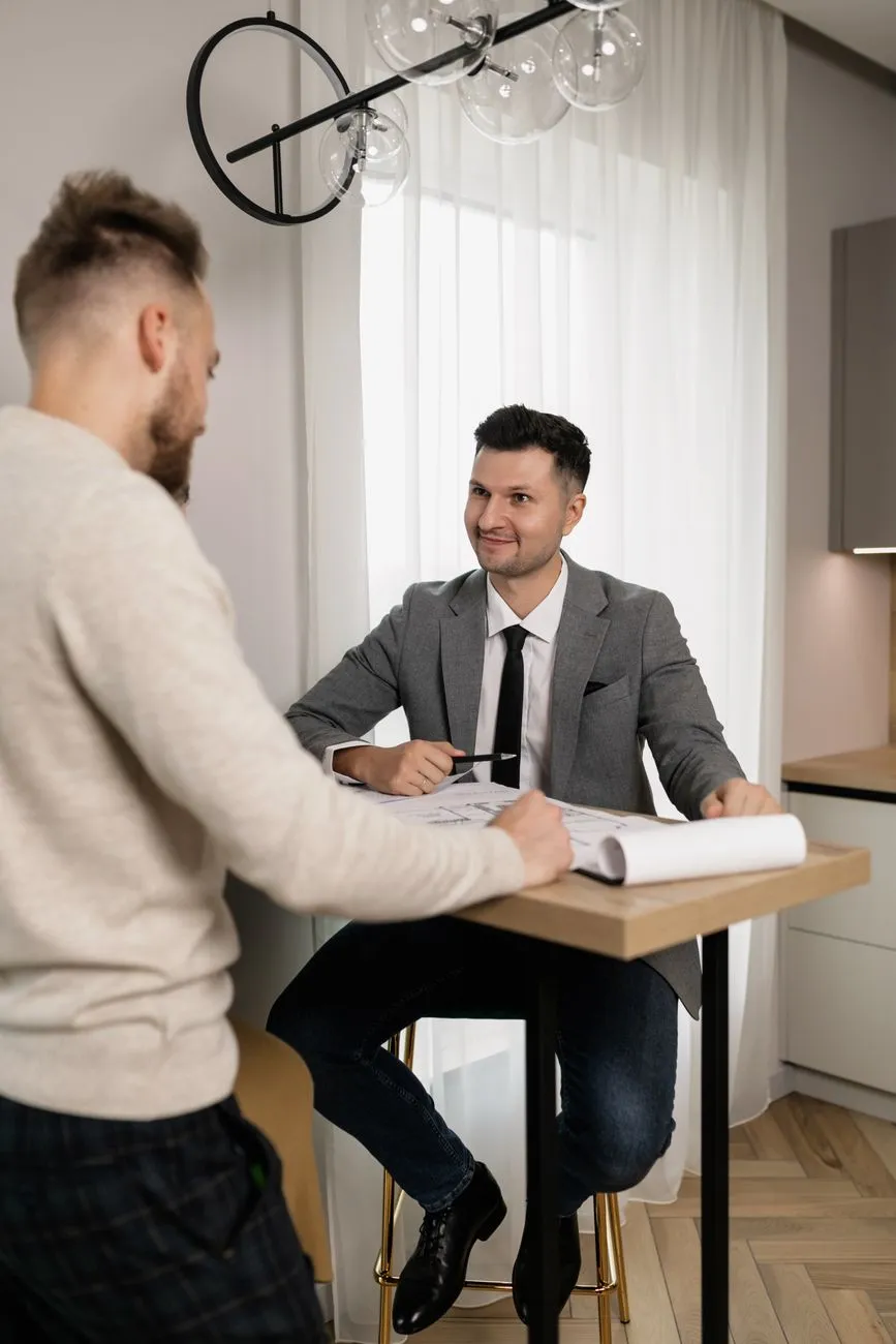 Zwei Männer, einer im Anzug, besprechen Pläne an einem Tisch in einem modernen Raum.