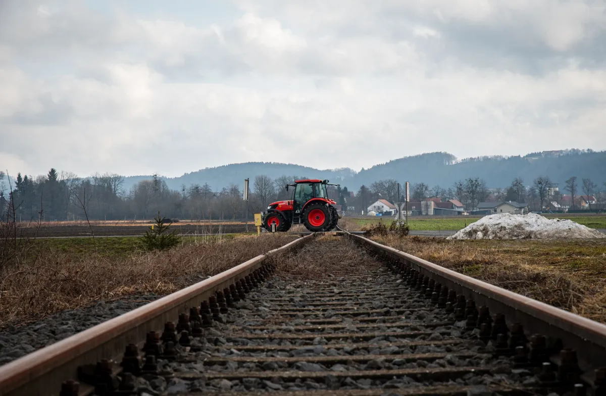 Traktor auf Bahngleisen in ländlicher Gegend mit Hügeln im Hintergrund und aufziehender Bewölkung.