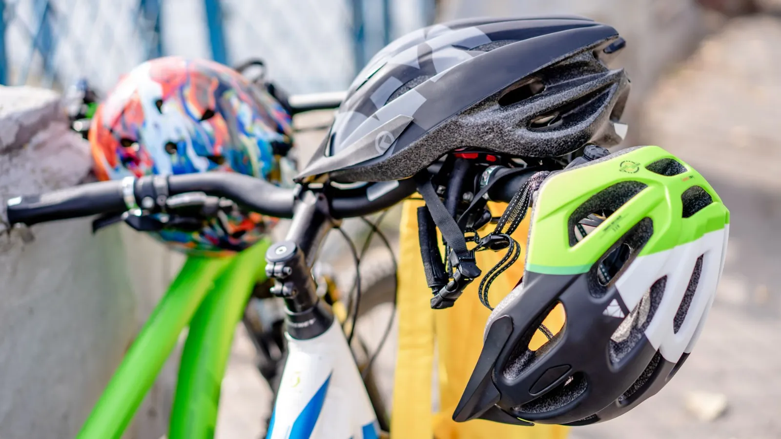 Fahrradhelme in verschiedenen Farben, darunter ein bunter, ein schwarzer und ein neongrüner Helm, hängen am Lenker eines Fahrrads.