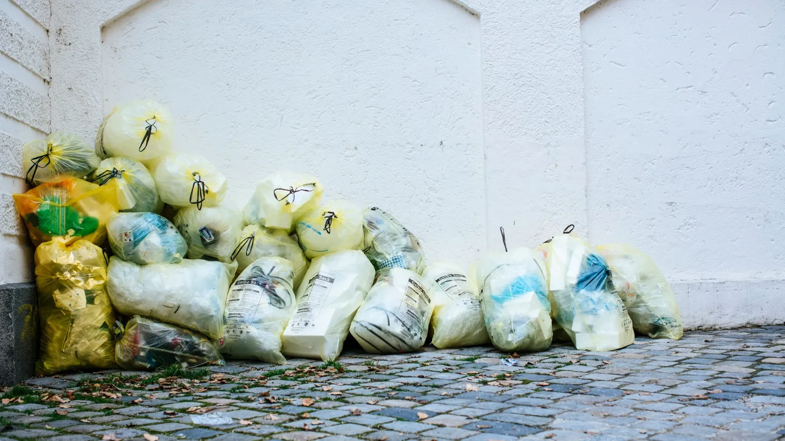 Gelbe Säcke mit Müll stapeln sich vor einer weißen Wand auf einem gepflasterten Gehweg.