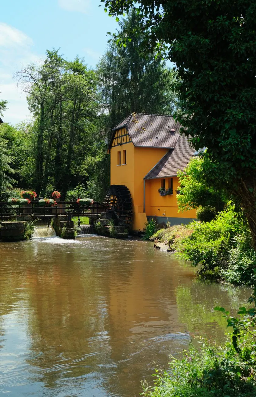 Wassermühle mit Wasserrad und Brücke mit Blumen, umgeben von Bäumen und Fluss.