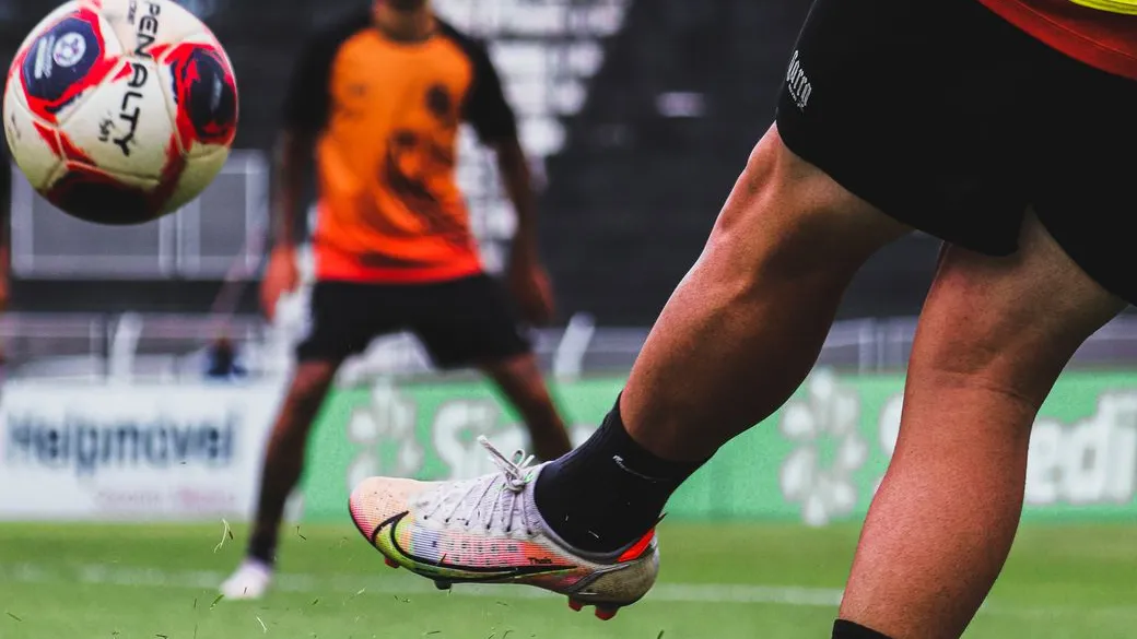 Fußballer tritt Ball am Rasen, Spieler in orange-schwarzem Trikot im Hintergrund