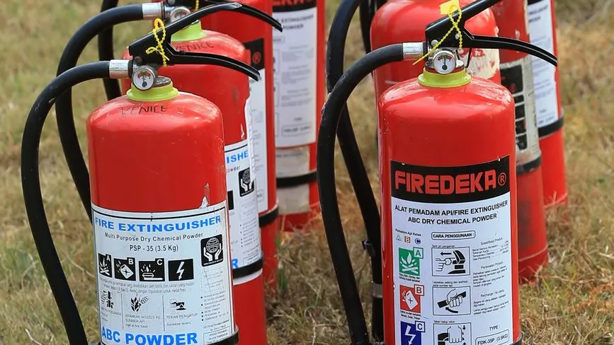 Feuerlöscher in Reihe auf Gras, rote Geräte mit schwarzen Schläuchen und Hinweisschildern.